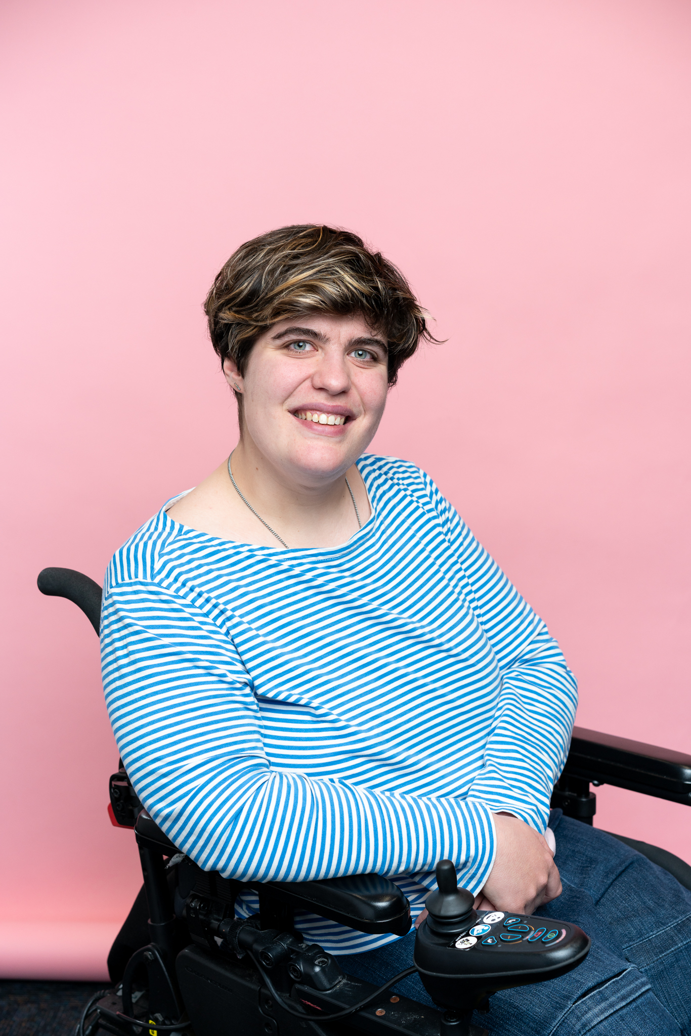 Henrietta (Etta) Bollinger, a person with short brown and blonde hair, smiles while seated in a power wheelchair against a plain pink background. They wear a blue and white striped long-sleeve top and blue jeans.